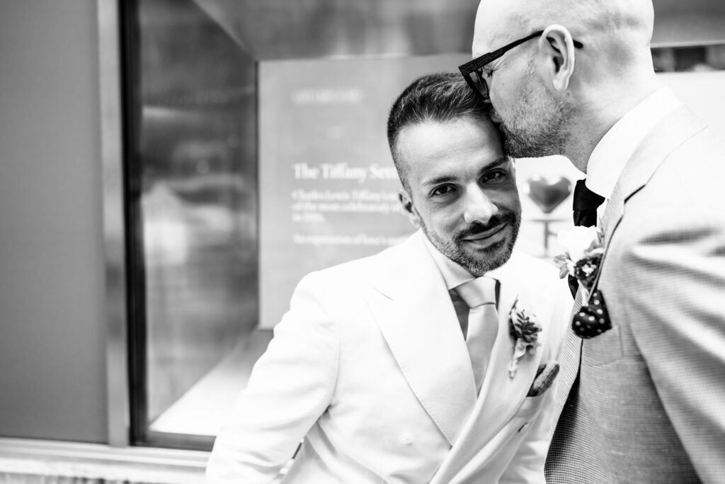 Two grooms sharing a kiss on NYC street with iconic buildings behind them, officiated by Jackie Reinking